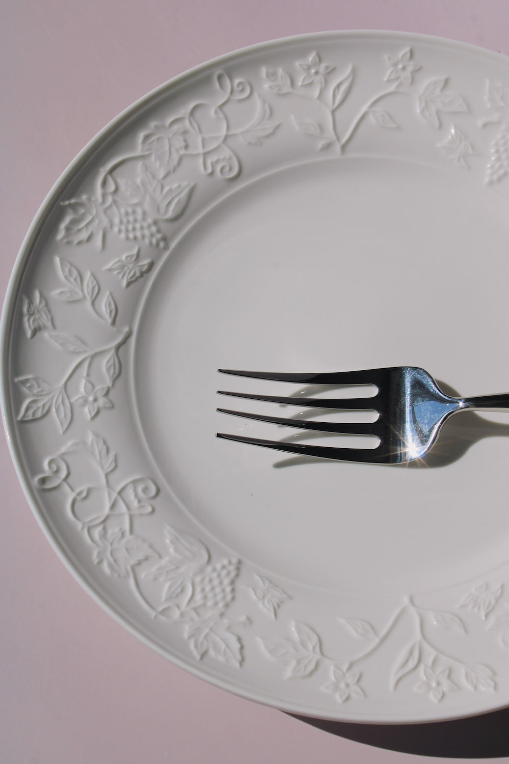 Florentia Ceramic Plate in white with embossed floral and botanical border detail, styled with a silver fork, on a soft pink background.