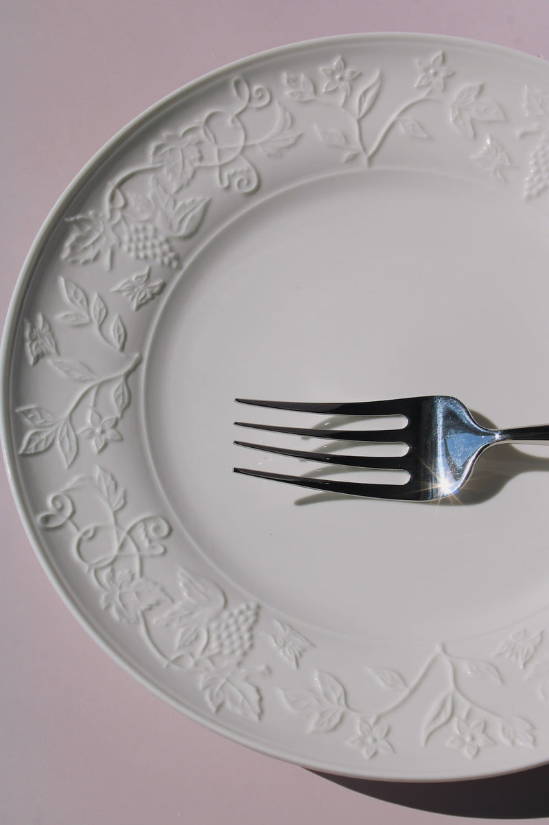 Florentia Ceramic Plate in white with embossed floral and botanical border detail, styled with a silver fork, on a soft pink background.
