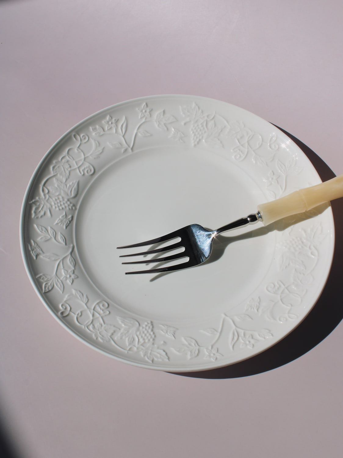 Florentia Ceramic Plate in white with embossed floral and botanical border detail, styled with a silver fork, on a soft pink background.