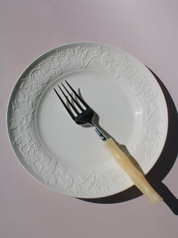 Florentia Ceramic Plate in white with embossed floral and botanical border detail, styled with a silver fork, on a soft pink background.