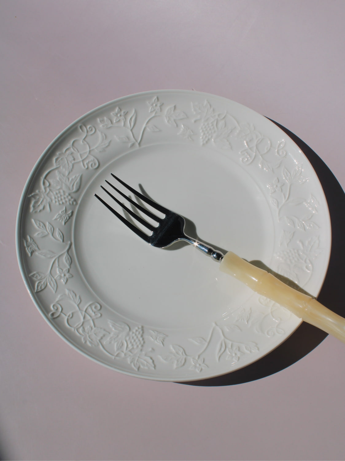 Florentia Ceramic Plate in white with embossed floral and botanical border detail, styled with a silver fork, on a soft pink background.