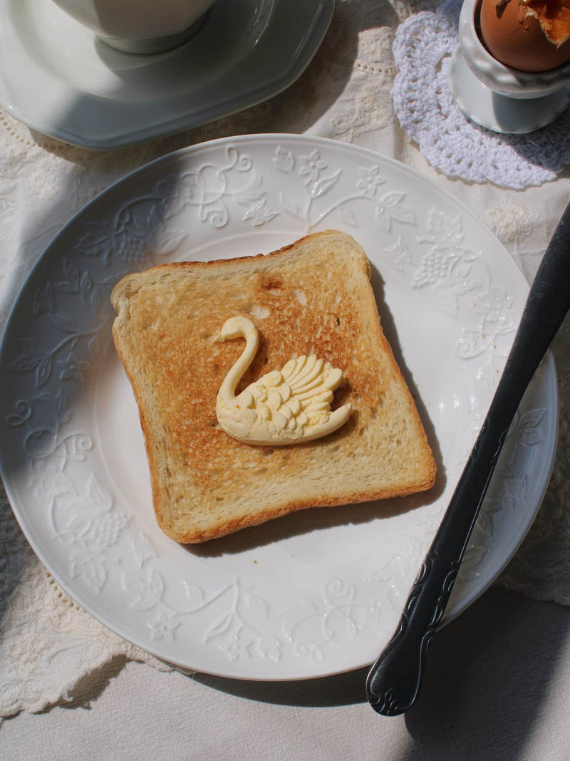 A square slice of toasted bread on a white embossed plate, topped with a pale yellow butter or cream swan shape, with a spoon and part of a cup visible in a styled tabletop setting.