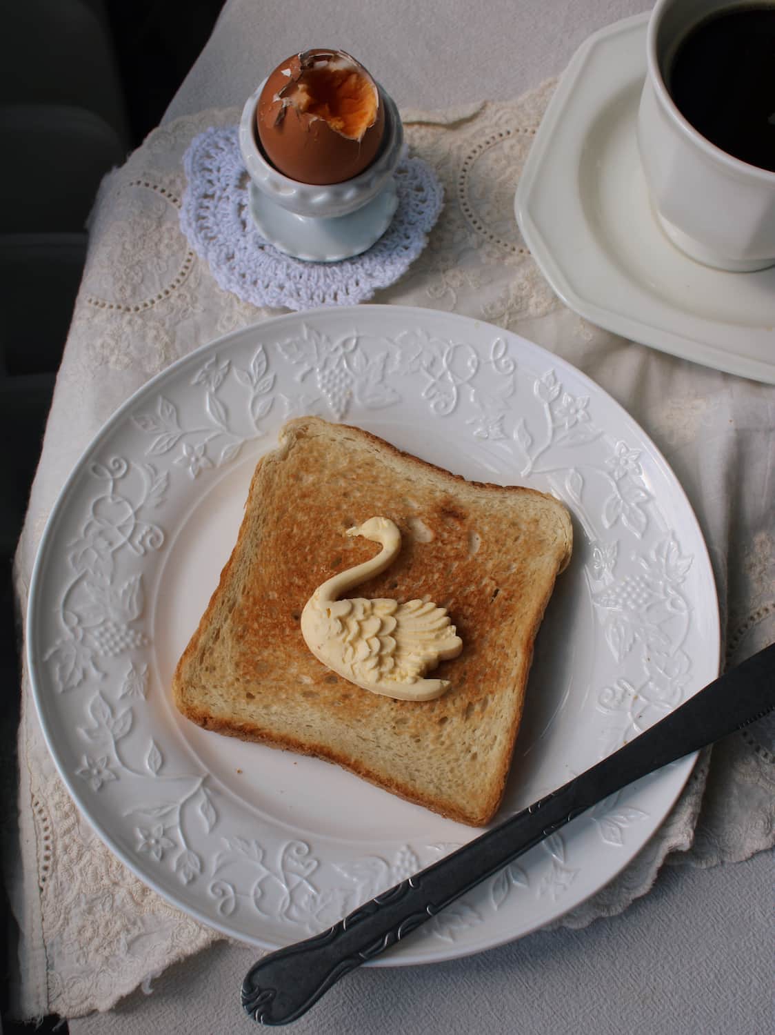 A slice of toasted bread on a white decorative plate, topped with a swan-shaped butter or pastry decoration, with a spoon and a cup of coffee on a lace tablecloth.