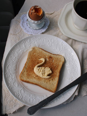 A slice of toasted bread on a white decorative plate, topped with a swan-shaped butter or pastry decoration, with a spoon and a cup of coffee on a lace tablecloth.
