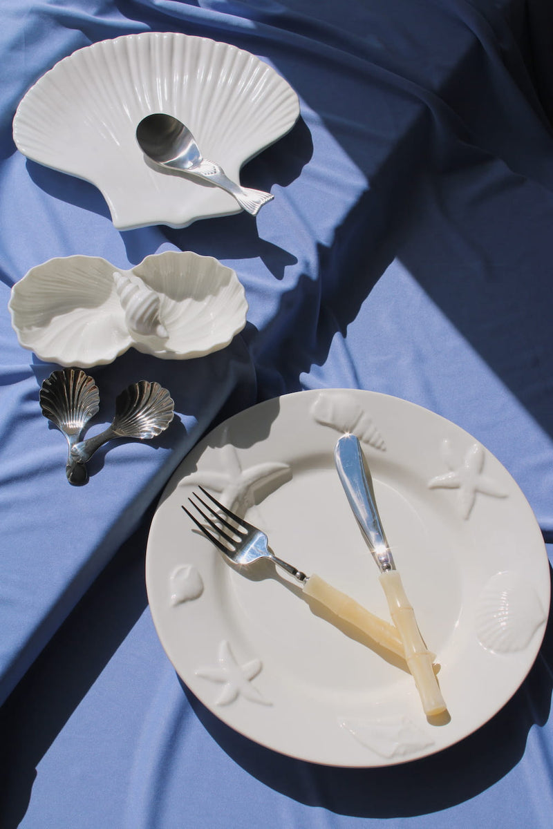 Caspia Ceramic Plate with starfish and shell relief detail, Calista Ceramic Shell Plate and Littora Ceramic Shell Dish styled together with silver shell-shaped spoons on a blue fabric surface in natural sunlight.
