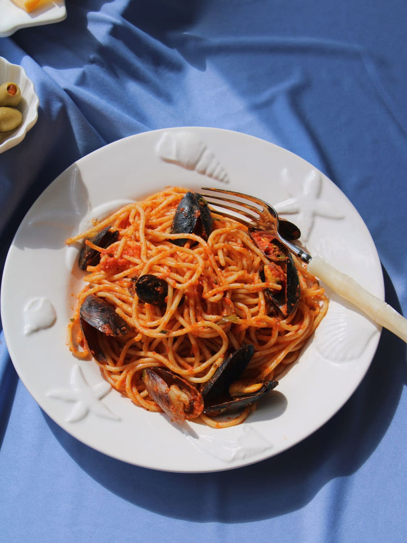Caspia Ceramic Plate in white with sculpted starfish and shell border, styled with spaghetti alle cozze and a fork on a blue fabric surface, with the Littora Ceramic Shell Dish with olives visible in the background.