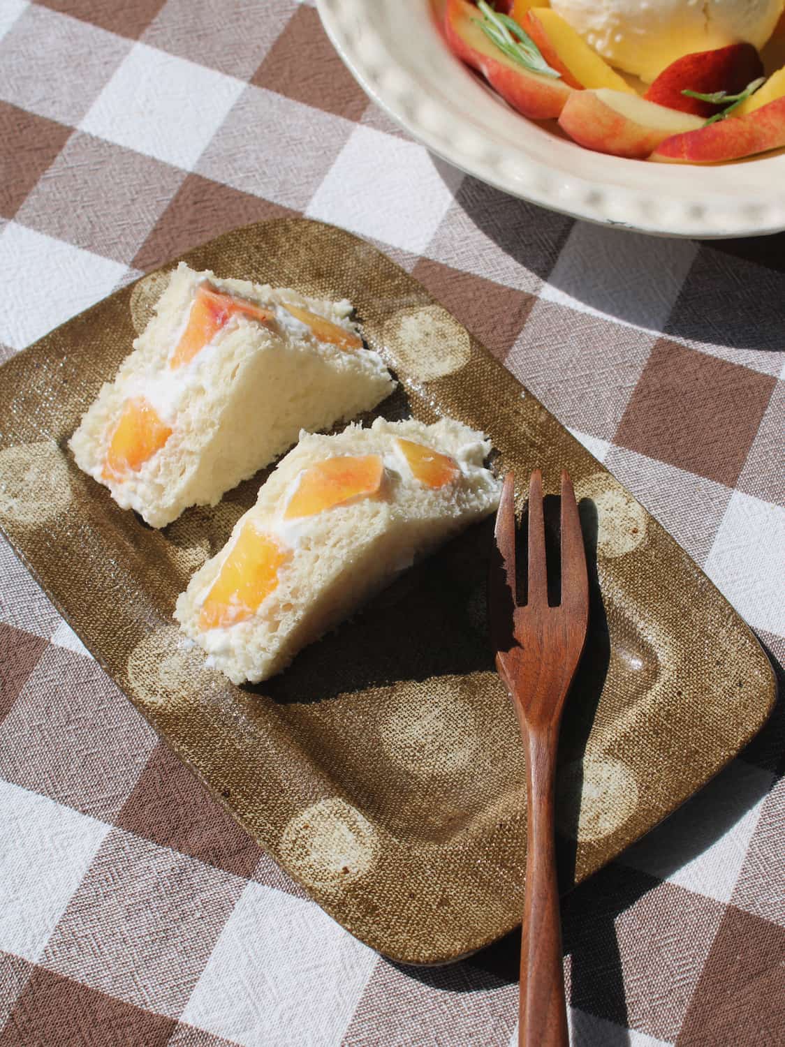 Three triangular sandwich cakes with peach slices on a textured Biscotti brown plate, set on a brown and white checkered tablecloth next to a wooden fork.