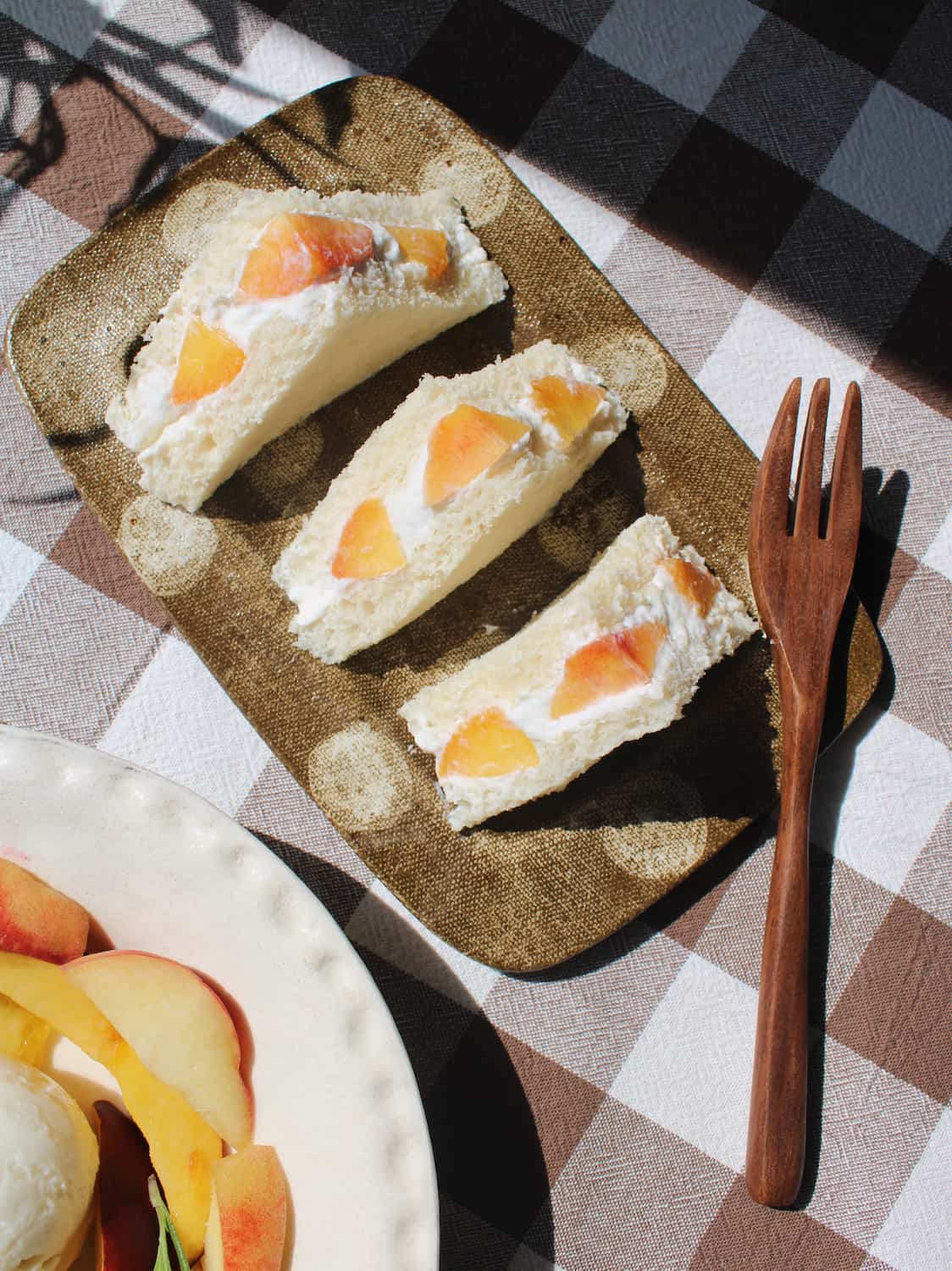 Three triangular sandwich cakes with peach slices on a textured Biscotti brown plate, set on a brown and white checkered tablecloth next to a wooden fork.