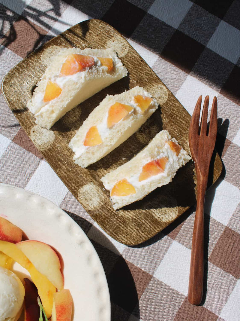 Three triangular sandwich cakes with peach slices on a textured Biscotti brown plate, set on a brown and white checkered tablecloth next to a wooden fork.