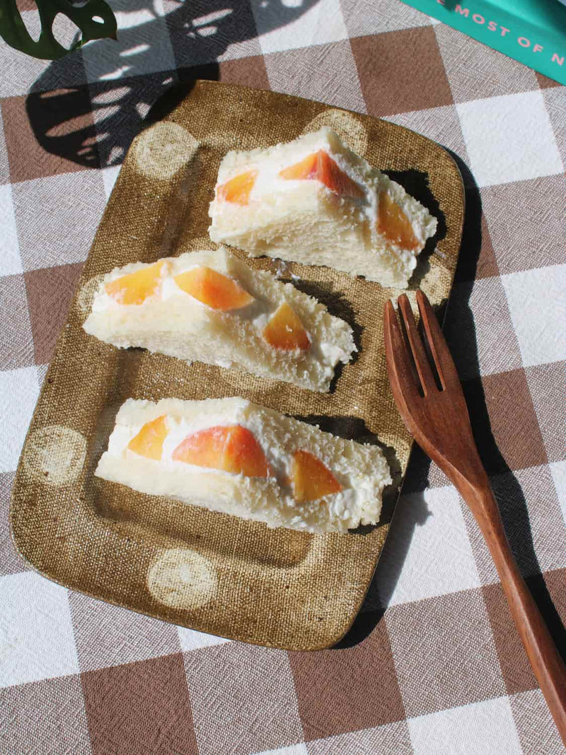Three triangular sandwich cakes with peach slices on a textured Biscotti brown plate, set on a brown and white checkered tablecloth next to a wooden fork.
