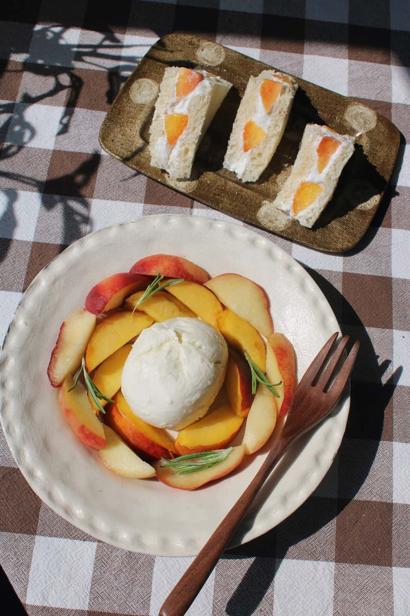 Three triangular sandwich cakes with peach slices on a textured Biscotti brown plate, set on a brown and white checkered tablecloth next to a wooden fork.