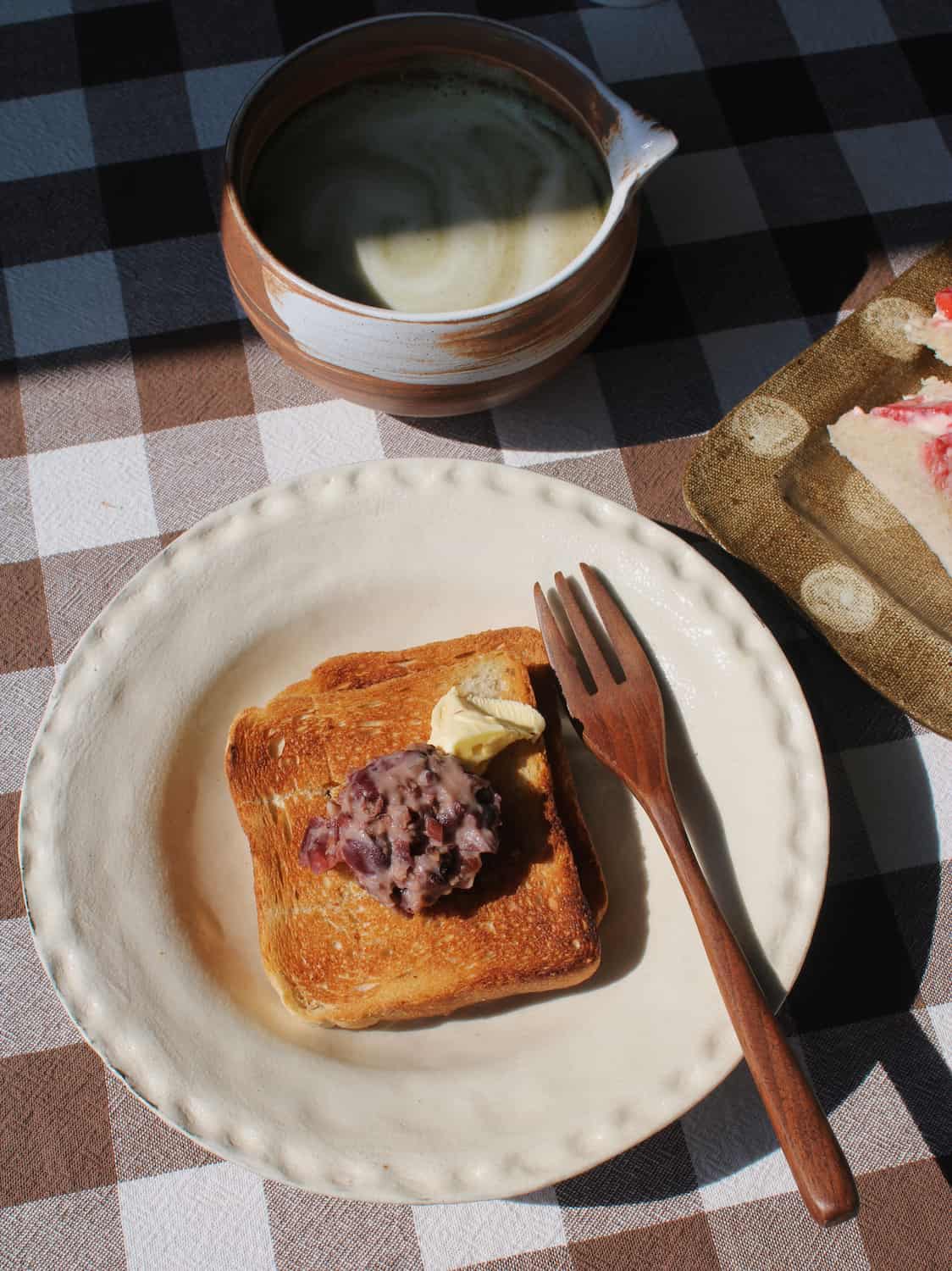 Lunette Ceramic Plate in cream with subtle textured scallop rim, styled with toast topped with red bean paste and butter alongside a wooden fork, with the Kanso Matcha Bowl and a silver ice cream dish visible in the background on a brown gingham surface