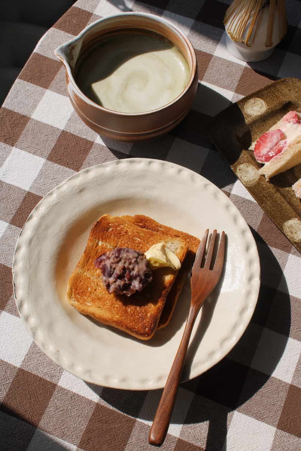 Lunette Ceramic Plate in cream with subtle textured scallop rim, styled with toast topped with red bean paste and butter alongside a wooden fork, with the Kanso Matcha Bowl and a silver ice cream dish visible in the background on a brown gingham surface