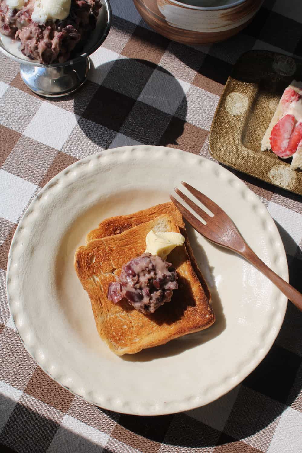 Lunette Ceramic Plate in cream with subtle textured scallop rim, styled with toast topped with red bean paste and butter alongside a wooden fork, with the Kanso Matcha Bowl and a silver ice cream dish visible in the background on a brown gingham surface