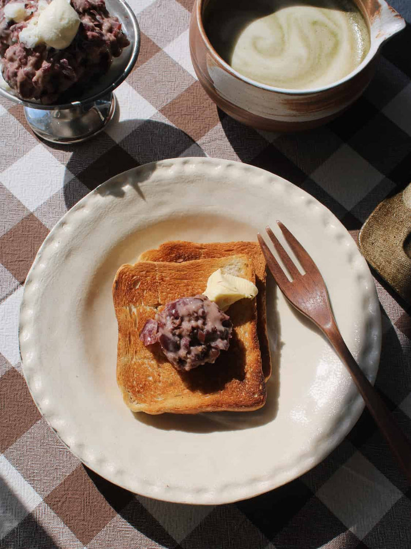 Lunette Ceramic Plate in cream with subtle textured scallop rim, styled with toast topped with red bean paste and butter alongside a wooden fork, with the Kanso Matcha Bowl and a silver ice cream dish visible in the background on a brown gingham surface