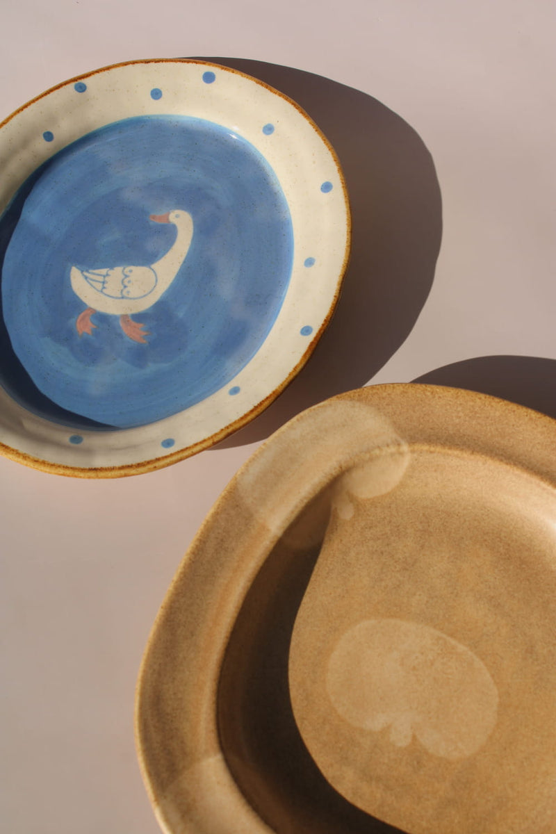 Blue ceramic plate with duck illustration and dotted rim beside a brown handmade ceramic plate, aesthetic tableware styling.