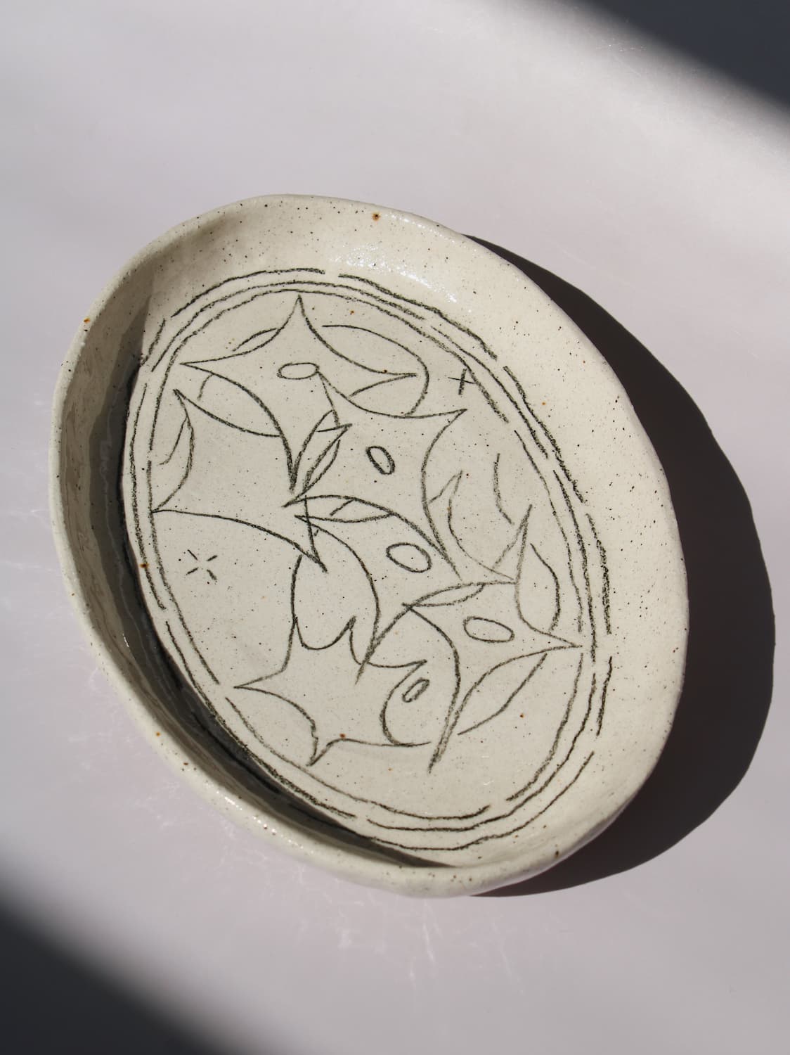 Mythos Ceramic Plate in speckled cream with hand-etched star and leaf motifs, viewed from above on a soft white background.