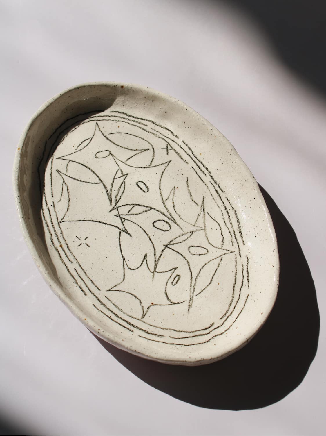Mythos Ceramic Plate in speckled cream with hand-etched star and leaf motifs, viewed from above on a soft white background.