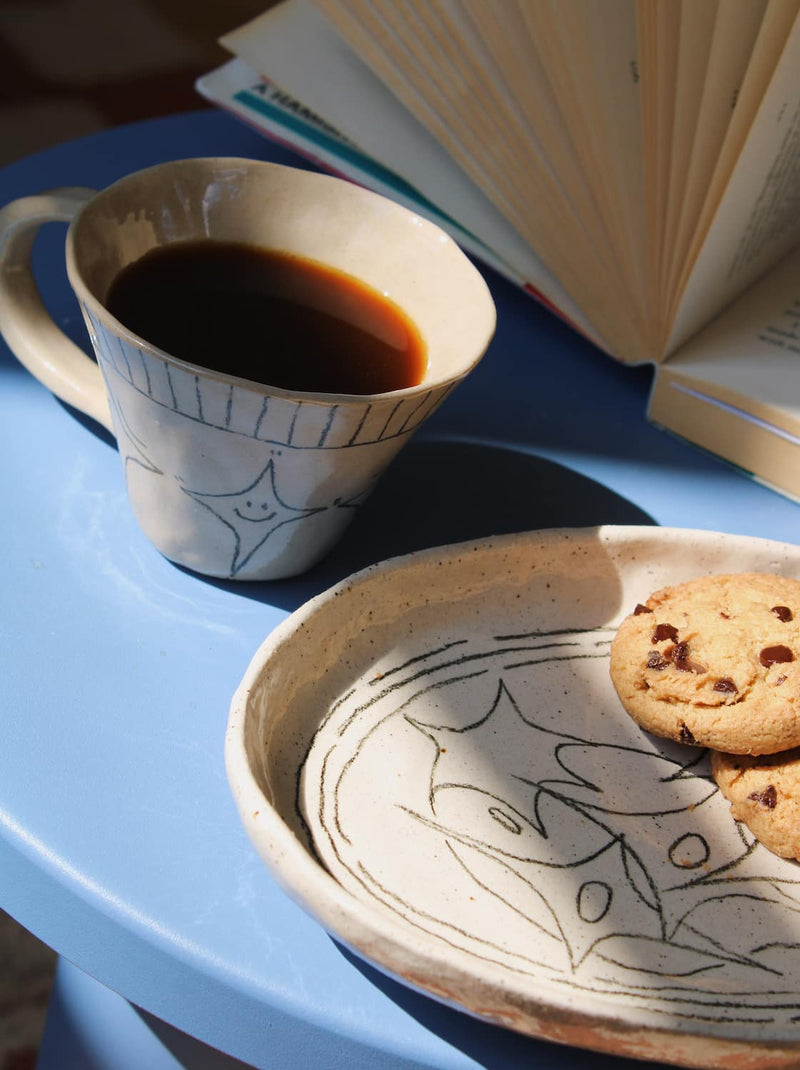 Astral Ceramic Mug in speckled cream with hand-etched star and stripe detail, held in hand filled with a latte, with a blurred indoor background in natural light.