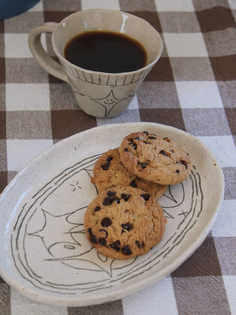 Mythos Ceramic Plate with hand-etched constellation detail styled with chocolate chip cookies alongside the Astral Ceramic Mug filled with black coffee, on a blue surface with an open book in natural sunlight.