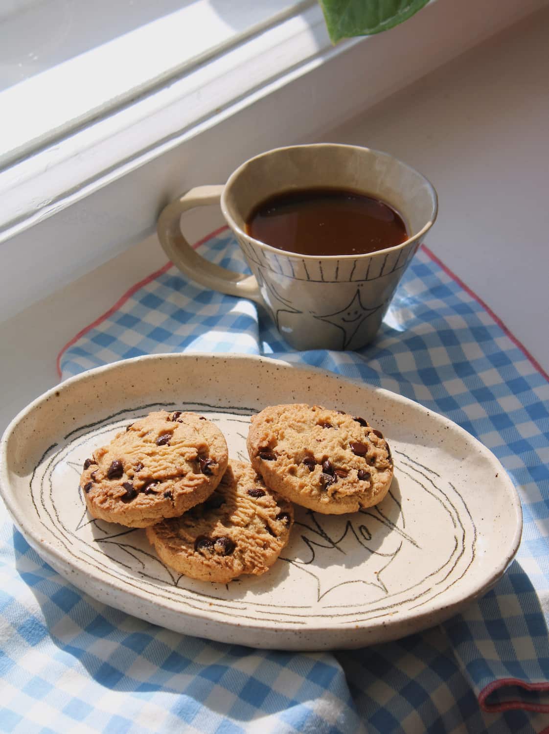 Mythos Ceramic Plate with hand-etched constellation detail styled with chocolate chip cookies alongside the Astral Ceramic Mug filled with black coffee, on a blue surface with an open book in natural sunlight.
