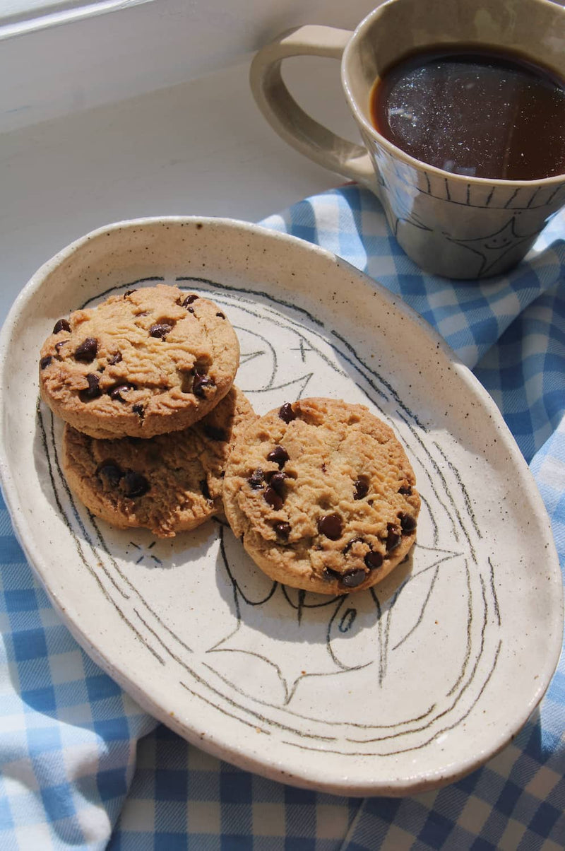Mythos Ceramic Plate in speckled cream with hand-etched star and constellation linework, styled with chocolate chip cookies on a blue surface with an open book in the background.