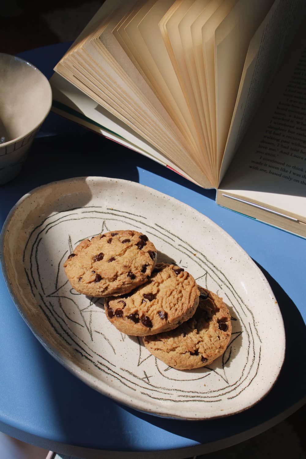 Mythos Ceramic Plate in speckled cream with hand-etched star and constellation linework, styled with chocolate chip cookies on a blue surface with an open book in the background.