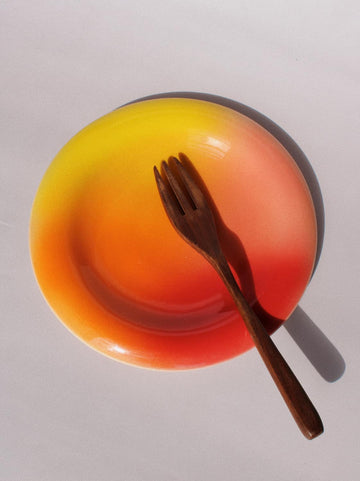 A ceramic plate with a vibrant yellow-to-orange-to-red gradient, topped with a dark brown wooden fork, set against a plain light gray background with a hard shadow.