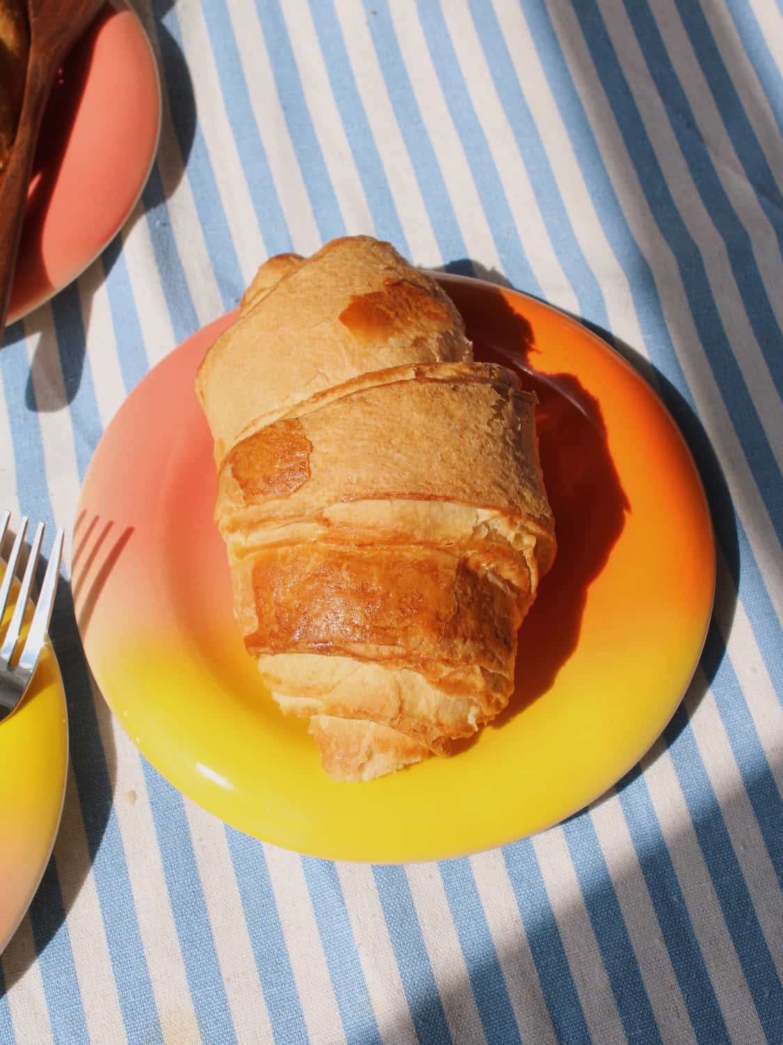 Belly Sunset Ceramic Plate by Agata Puławska – colourful handmade ceramic plate styled with a croissant on a blue striped tablecloth