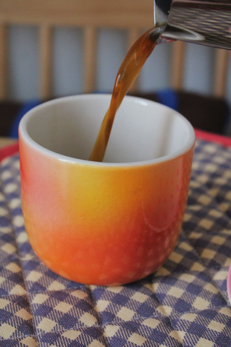Sunset Gradient Ceramic Cup by Agata Puławska – colourful handmade ceramic cups styled with coffee and a pastry on a blue striped tablecloth
