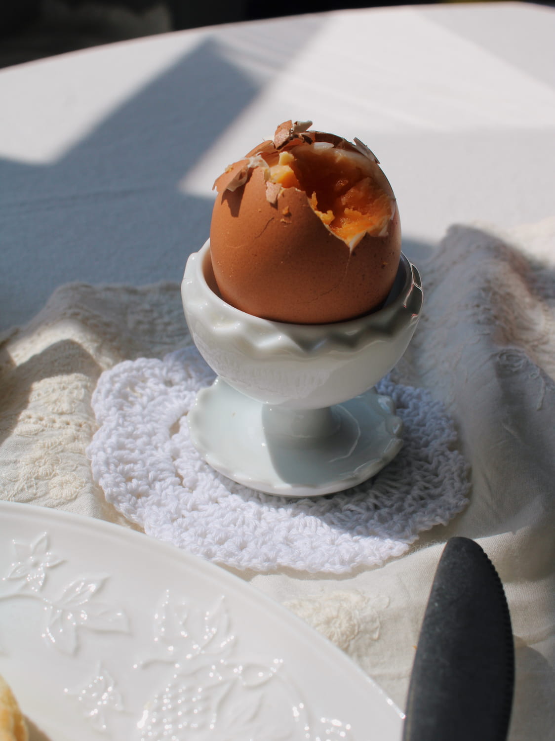 A cooked brown egg standing upright in a white ceramic egg cup, placed on a white lace doily on a white tablecloth with strong sunlight and shadows.