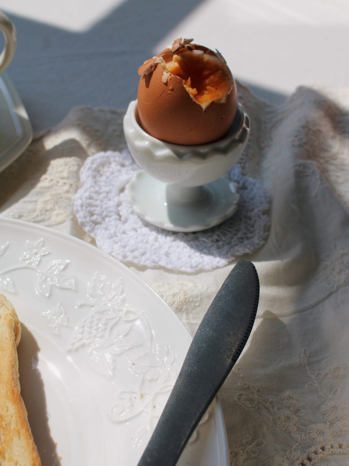 A hard-boiled egg with a cracked shell sits in a white ceramic egg cup on a lace doily, surrounded by a white plate, butter knife, and cream tablecloth in bright natural light.