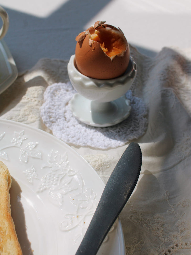A hard-boiled egg with a cracked shell sits in a white ceramic egg cup on a lace doily, surrounded by a white plate, butter knife, and cream tablecloth in bright natural light.