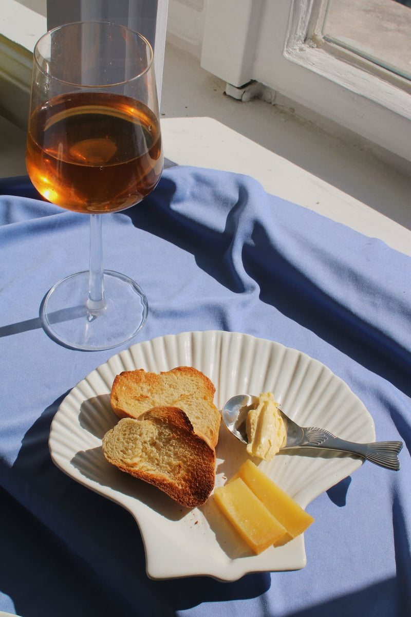 Calista Ceramic Shell Plate styled with toasted bread, cheese and butter alongside the Pisca Spoon, with a glass of rosé wine on a blue fabric surface by a sunlit window.