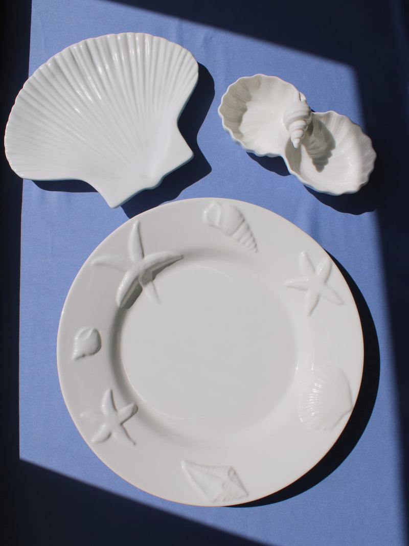 Calista Ceramic Shell Plate, Littora Ceramic Shell Dish and Caspia Ceramic Plate displayed together in white ceramic on a blue fabric surface in natural sunlight, showing the full coastal collection.