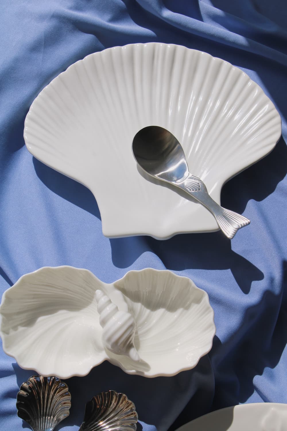 Calista Ceramic Shell Plate, Littora Ceramic Shell Dish displayed together in white ceramic on a blue fabric surface in natural sunlight, showing the full coastal collection