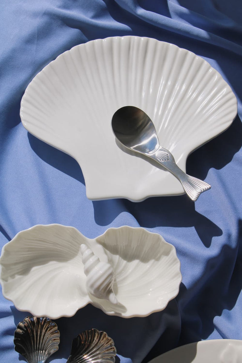 Calista Ceramic Shell Plate, Littora Ceramic Shell Dish displayed together in white ceramic on a blue fabric surface in natural sunlight, showing the full coastal collection