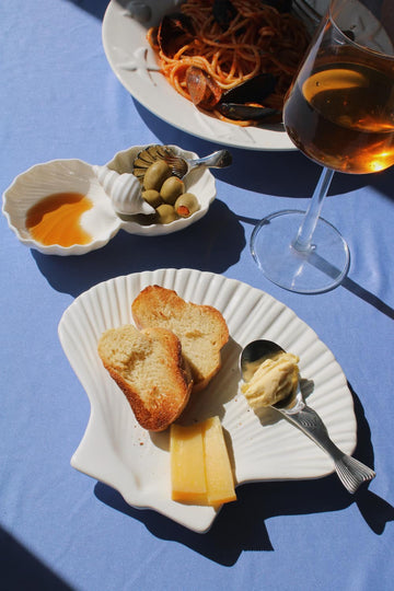 Littora Ceramic Shell Dish filled with olive oil and olives alongside the Calista Ceramic Shell Plate styled with toasted bread, cheese and butter, with Venera Shell Spoons and Pisca Spoon, a glass of rosé wine and the Caspia Ceramic Plate with spaghetti alle cozze visible in the background on a blue fabric surface.