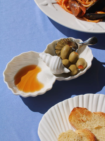 Littora Ceramic Shell Dish in white with two scallop-shaped compartments, styled with Venera Shell Spoons on a blue fabric surface