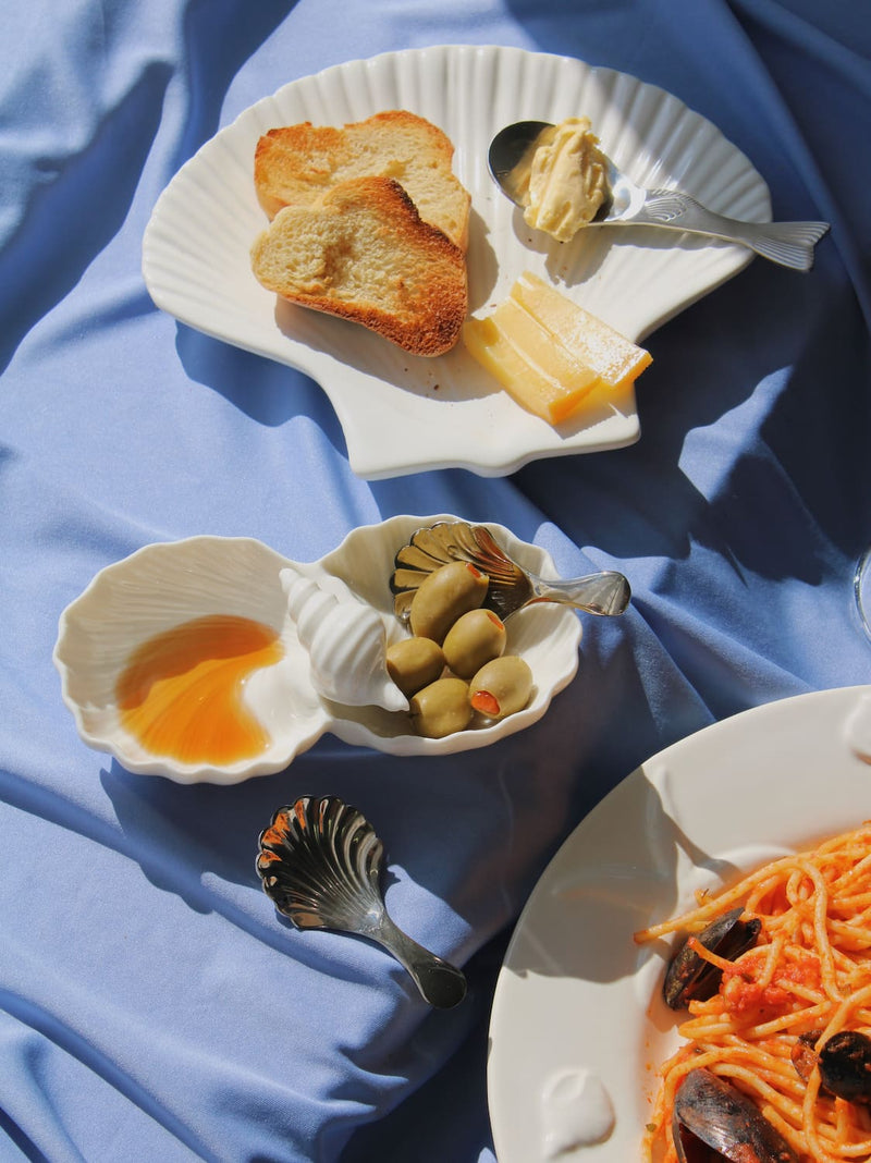 Littora Ceramic Shell Dish filled with olive oil and olives alongside the Calista Ceramic Shell Plate styled with toasted bread, cheese and butter, with Venera Shell Spoons and Pisca Spoon, a glass of rosé wine and the Caspia Ceramic Plate with spaghetti alle cozze visible in the background on a blue fabric surface.