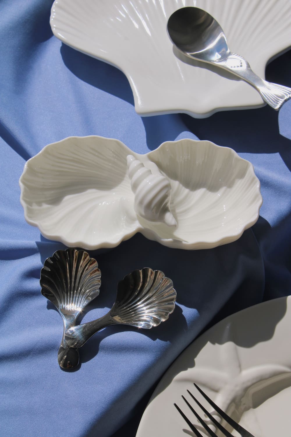 Littora Ceramic Shell Dish in white with two scallop-shaped compartments, styled with Venera Shell Spoons on a blue fabric surface