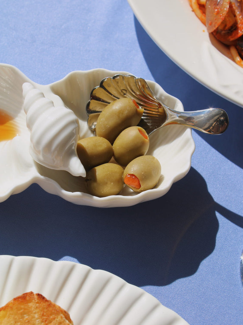 Littora Ceramic Shell Dish in white with two scallop-shaped compartments, styled with Venera Shell Spoons on a blue fabric surface