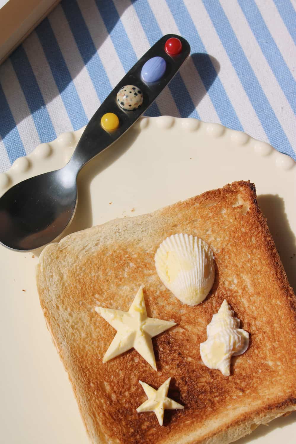 A styled food scene showing a slice of toasted bread on a cream plate, decorated with three small seashells and two star-shaped butter pieces. A spoon with colorful shell-like accents on the handle rests on a blue-and-white striped cloth in the background.