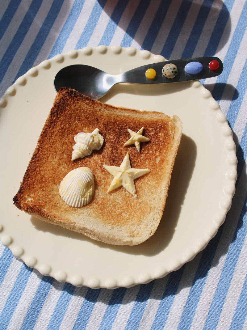A square slice of toasted bread on a white scalloped plate, decorated with three seashell-shaped butter puffs and two star-shaped butter puffs. A spoon with colorful decorative beads on the handle rests on the plate, all set on a blue and white striped fabric background.