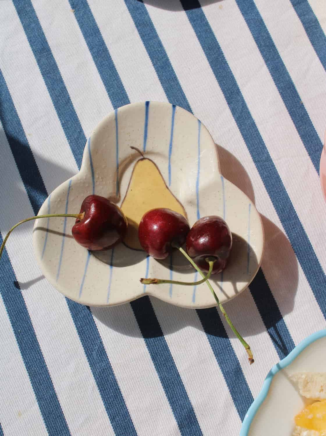 A small ceramic dish with a scalloped edge and thin blue stripes, holding three red cherries and a pale yellow pear on a blue-and-white striped tablecloth.