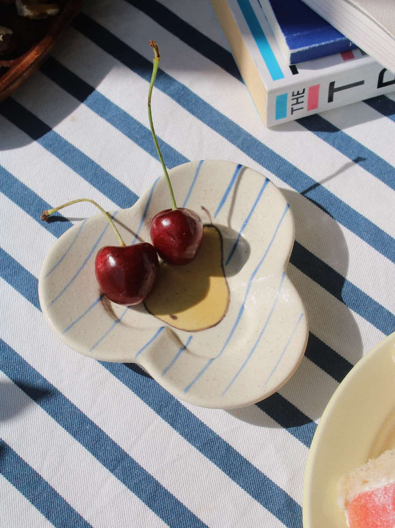 A small cream-colored ceramic dish with blue stripes and a flower-like shape, holding two red cherries on a blue and white striped tablecloth.