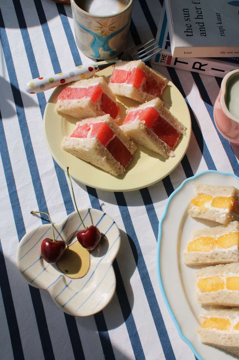 A styled tabletop scene with sliced sandwich cakes on plates, two cherries on a small dish, a striped tablecloth, books, and mugs in warm natural light.