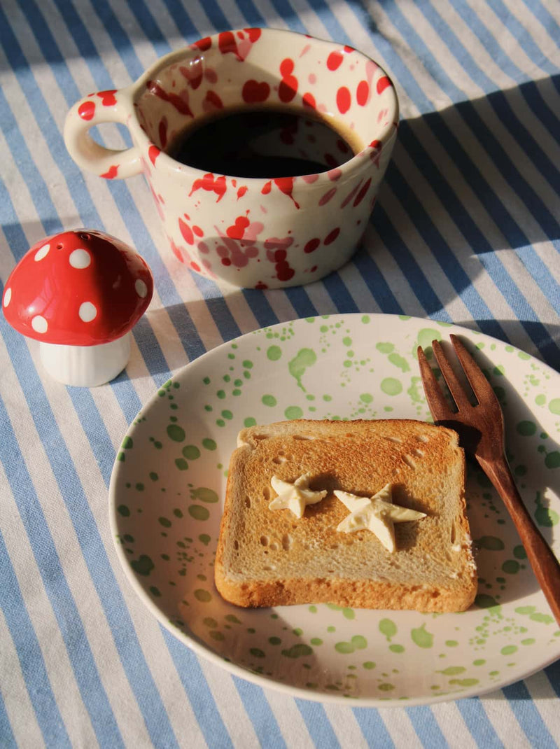A styled breakfast scene on a blue and white striped tablecloth. A white ceramic mug with red polka dots sits filled with black coffee, next to a small red-and-white mushroom-shaped container.