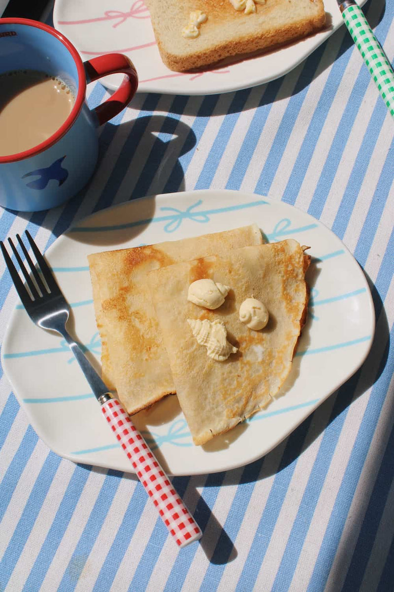 A styled breakfast scene on a blue and white striped tablecloth. In the foreground, a white plate holds two folded crepes topped with butter and a fork with a red-and-white gingham handle.