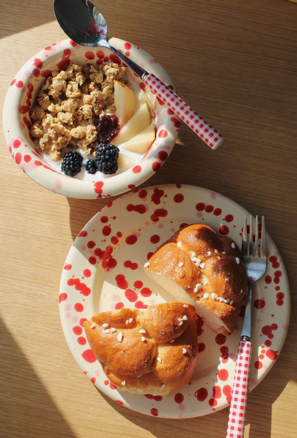 Two speckled ceramic bowls and plates on a wooden table, styled with breakfast food. One bowl contains granola with fruit and berries, and the other holds sliced bread with sea salt.